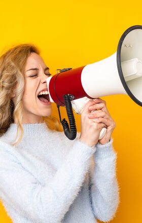 Eine Frau steht vor einem gelben Hintergrund, sie hält ein Megafon in der hand in welches Sie hinein schreit.