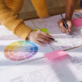 Foto eines Schreibtisches auf welchem Arbeitspapiere liegen, die Papiere zeigen Skizzen des Aufbaus einer Website sowie einen Farbkreis. Man sieht die Hände zweier Personen welche Stifte in der Hand halten und an den Papieren arbeiten.