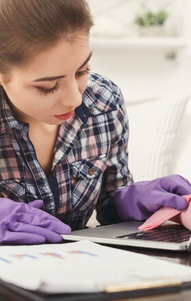 Eine Frau Reinigt einen Laptop mit einem Putzlappen, sie trägt dabei Gummihandschuhe.